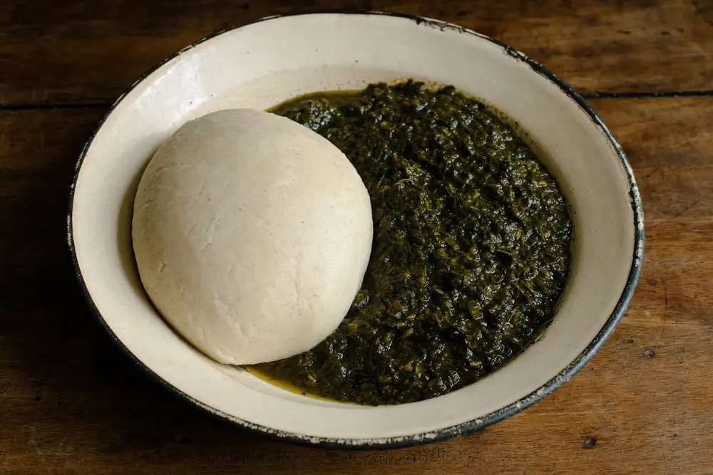 Une boule de foutou pilé, beige clair et mate, à côté d’une sauce feuille vert sombre dans un plat émaillé.