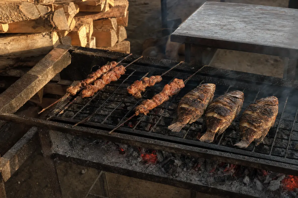 Un gril de plein air au feu de bois, brochettes et poissons posés sur la grille, fumée et bois fendu à côté.